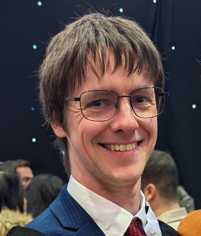 An image of Adam Hollings showing him smiling, wearing glasses. He has white skin, blue eyes, mousey blond hair and is wearing a blue suit jacket, red tie and white shirt.